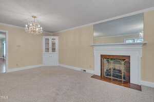 Carpeted living room with beige walls, a white fireplace with a large mirror above it, a brass fireplace screen, and a built-in white cabinet. A chandelier hangs from the ceiling.