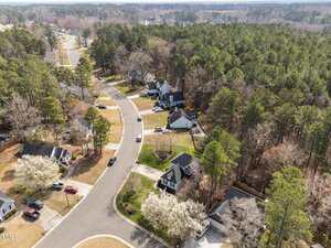 Aerial view of a suburban neighborhood with houses, driveways, cars, and tree-lined streets bordering a wooded area on a clear day.