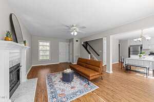 A modern living room with wood flooring, a brown leather sofa, a small ottoman on a patterned rug, a white fireplace, and an open view of the dining area and kitchen with stainless steel appliances.