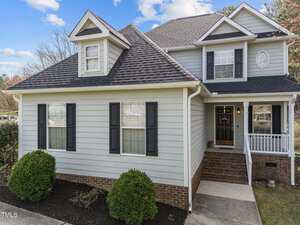 A two-story house with light gray siding, dark shutters, and a brick foundation. There are trimmed bushes in front, a small covered porch with white railings, and a dormer window under a blue sky.