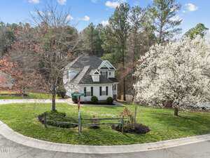 Two-story suburban house with a gray roof and white exterior on a corner lot, surrounded by green grass, blooming trees, and a wooden fence. A street sign and American flag are visible in the yard.