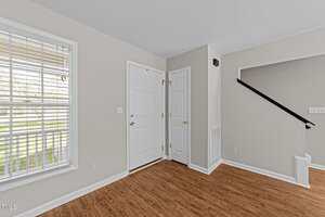 A bright entryway with light gray walls, a white front door, a small closet with a white door, brown wood flooring, a window with white blinds, and a black handrail leading up a staircase.