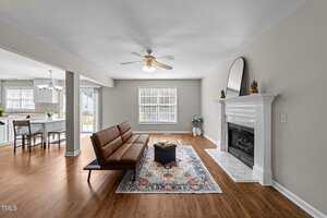 A living room with pale green walls, wood flooring, a brown sofa, a patterned rug, and a white fireplace with a round mirror and plants on the mantel. An open kitchen with dining area is visible in the background.