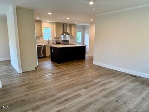 Open-concept room with light wood flooring, beige walls, and recessed lighting. A kitchen with stainless steel appliances, dark island, and white cabinets is visible in the background. Two windows provide natural light.