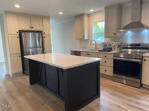 Modern kitchen with stainless steel appliances, light-colored cabinets, a black island with a light countertop, wood flooring, and a window above the sink. The room is well-lit with recessed ceiling lights.