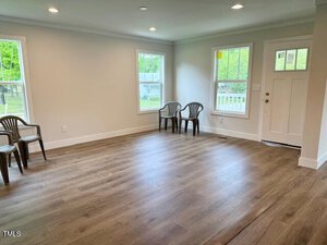 A mostly empty room with light-colored walls, wood flooring, three plastic chairs against the walls, two windows, and a white front door with a window. Natural light is coming in from the windows.