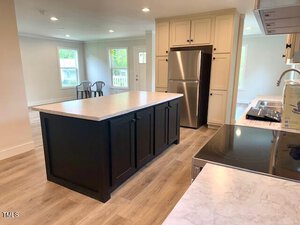 A modern kitchen with light wood floors, a large dark island, light cabinetry, stainless steel refrigerator, electric stovetop, and two chairs near windows in the background.