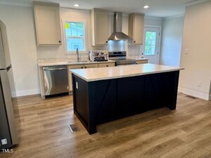 A modern kitchen with light-colored cabinets, stainless steel appliances, a center island with a dark base, marble countertops, wood flooring, and a window above the sink.