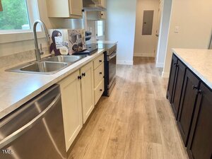 A modern kitchen with light wood flooring, white and dark cabinetry, stainless steel appliances, a double sink, and a countertop with a cookbook and utensils. A laundry or utility room is visible in the background.