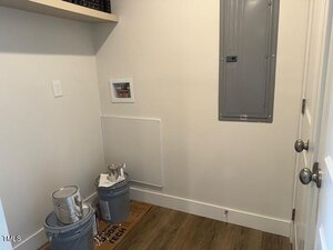 A small utility room with white walls, an electrical panel, a shelf, two paint cans, a paint tray, and a cardboard box on a wood floor. There is a cutout in the wall for laundry connections.