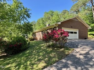 A single-story brick house with white garage doors, a sloped driveway, and a front yard featuring blooming red and pink bushes, surrounded by green trees under a clear blue sky.