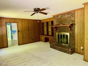 A living room with wood-paneled walls, beige carpet, a ceiling fan, a brick fireplace with a wooden mantel, built-in shelves and cabinets, and a doorway leading to a blue and white kitchen.
