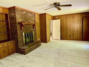 A living room with wood-paneled walls, a brick fireplace with a brass screen, built-in shelves and cabinets, white carpet, a ceiling fan, and an open door leading to another room.