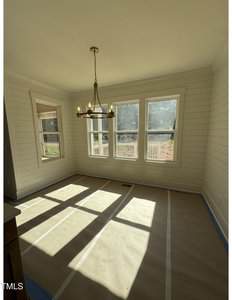 A bright room with three large windows, white shiplap walls, a modern brass chandelier, and the floor covered with protective paper lined in blue tape. Sunlight streams in, casting shadows on the floor.