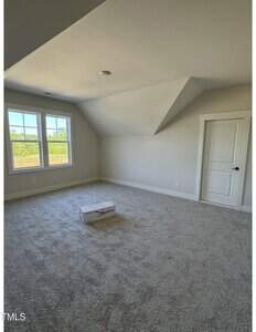 A carpeted, empty room with light-colored walls, a white door, two windows with outdoor views, and a small white box placed on the floor. The ceiling has an angled section.