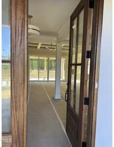 A wooden door is open, revealing an empty, unfinished room with covered floors, white walls, large windows, and modern light fixtures. Natural light enters from the windows, and the space appears to be under construction or renovation.