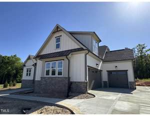 A two-story modern suburban house with white siding, stone accents, and a dark roof. The home features a double garage, multiple gables, and large windows. The driveway and yard are unfinished, with some construction materials visible.