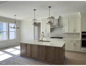 A modern kitchen with white cabinets, a large central island with a wood finish, gold pendant lights, a built-in oven, gas cooktop, stainless steel hood, and large windows letting in natural light.
