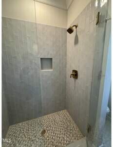 A walk-in shower with light gray rectangular wall tiles, a built-in wall niche, brass fixtures including a showerhead and handle, and a pebble-patterned tile floor with a circular drain.