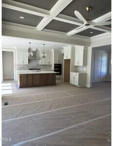 A modern kitchen with white cabinets, a center island with a wood finish, two pendant lights, and a coffered ceiling. The floor is covered with protective paper, indicating ongoing or recent construction.