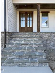 A set of wide stone steps leads up to a front porch with double wooden doors. The house exterior features light-colored siding, stonework, and a window to the right of the doors.