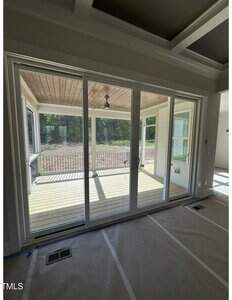 Sunlight enters through large glass sliding doors that lead from an indoor room with covered floors to a screened-in porch with a wooden ceiling, railing, and view of trees outside.