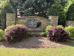 A stone wall with a sign reading "Silver Leaf - The Estates" stands at 1121 Dovefield Lane, Youngsville, surrounded by bushes and trees with sunlight casting shadows on the grass in front.