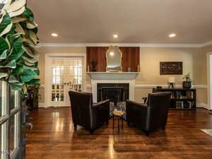 A living room with wooden floors, two black armchairs facing a fireplace with a mirror above it, shelves on the right, double French doors on the left, and a decorative wreath on the foreground door.