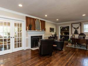 A living room with hardwood floors, two black armchairs, a fireplace with a mirror above it, built-in shelves, a wooden desk, and wall art featuring a deer head. French doors lead to another room.