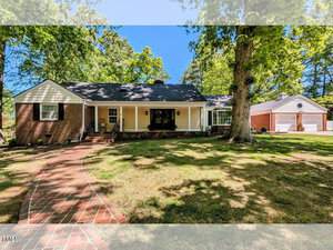 A single-story brick and siding house at 107 Forest Road, Oxford, features a covered front porch, large front window, and black roof on a shaded lot with mature trees and a curved brick walkway. Detached garage is visible to the right.