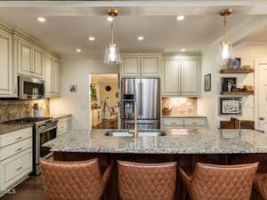 A modern kitchen features a large granite island with a built-in sink, three brown quilted barstools, stainless steel appliances, white cabinets, and pendant lights hanging from the ceiling.