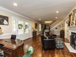 A spacious living room with hardwood floors, a wooden desk with a laptop, two black armchairs, a patterned ottoman, a fireplace, and a staircase leading to the upper floor. Large windows let in natural light.