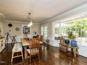 A dining room with wooden floor, a rectangular table with mixed wood chairs and benches, a large window on the right, shelves with decorative items, and various potted plants along the window.