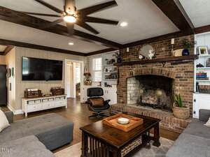 A cozy living room features a large brick fireplace with a wood mantel, built-in shelves, a wall-mounted TV, modern gray sectional sofas, a wooden coffee table, and a black lounge chair on dark hardwood floors.