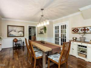 A dining room with a wooden table, six chairs, a brown table runner, white walls with wainscoting, hardwood floors, a glass light fixture, and a sideboard with decor. French doors and a large framed painting are visible.