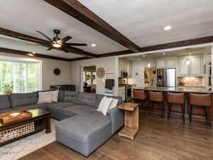 A living room with a gray sectional sofa, wooden coffee table, and side table is adjacent to a kitchen with a breakfast bar, four brown barstools, white cabinets, and stainless steel appliances. Exposed beams cross the ceiling.