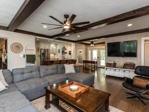 A spacious living room with a gray sectional sofa, wooden coffee table, and TV mounted on a paneled wall. The area has exposed wooden ceiling beams, ceiling fans, and an open view into the kitchen and dining area.