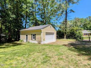 A detached yellow garage with white trim sits on a grassy yard, surrounded by tall trees and a black fence, with a neighboring house visible in the background.