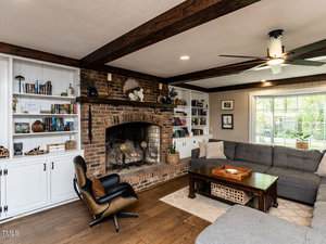A living room with a brick fireplace, built-in white shelves filled with books and decor, a gray sectional sofa, a wooden coffee table, hardwood floors, and ceiling beams. A large window lets in natural light.