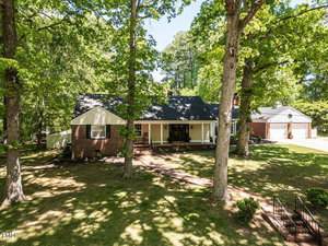 Nestled in Oxford, this single-story brick house at 107 Forest Road sits among large mature trees. The shaded front yard and driveway lead to a detached garage, while sunlight filters through the leafy canopy onto the lawn.