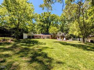 Single-story house with a large, grassy backyard surrounded by trees. The house has multiple sections and a patio with outdoor seating. The area is shaded by mature trees and bathed in sunlight.