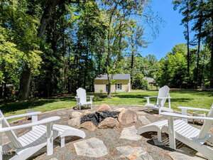 Four white Adirondack chairs surround a circular stone fire pit on a gravel area in a grassy backyard with tall trees and a yellow house in the background.