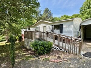 A white single-story house at 328 Cone Drive, Henderson features a wooden wheelchair-accessible ramp, lattice skirting, and a carport. Surrounded by trees and greenery, the sunny yard includes shrubs and gravel.