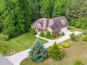 Aerial view of a two-story brown house at 1619 Carriage Drive, Franklinton, with a two-car garage, well-maintained lawn, bushes, and tall trees. A curved driveway leads from the street to the garage and front entrance.