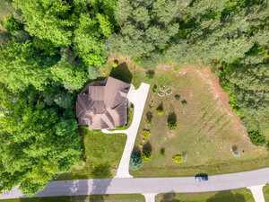Aerial view of a house with a beige roof and curved driveway surrounded by trees and a large grassy yard; a single car is parked on the street in front of the property.