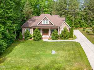 A single-story house with a brown roof and brick exterior sits on a well-kept lawn, surrounded by trees. A concrete driveway runs along the right side of the house, and shrubs line the front porch.