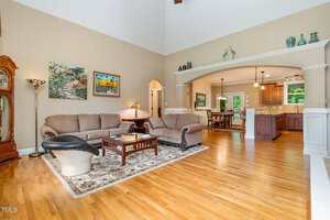 A spacious living room with hardwood floors, beige walls, two matching sofas, a patterned area rug, a wooden coffee table, and an open view into a kitchen and dining area with wood cabinetry and bar seating.