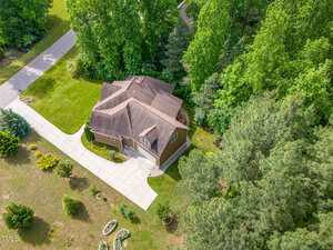 Aerial view of a two-story house with a brown roof and attached garage, surrounded by a large green lawn, trees, and landscaping. A long driveway curves from the street to the garage. Dense woods border the property.