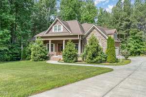 A two-story house with brown siding and stone accents, a covered front porch with columns, surrounded by trees and shrubs, set on a neatly manicured lawn with a curved concrete driveway.