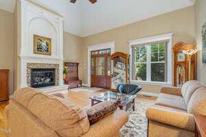 A living room with beige walls, a high ceiling, a fireplace with a white mantel, a brown sofa and loveseat, a glass coffee table, an area rug, and large windows letting in natural light.